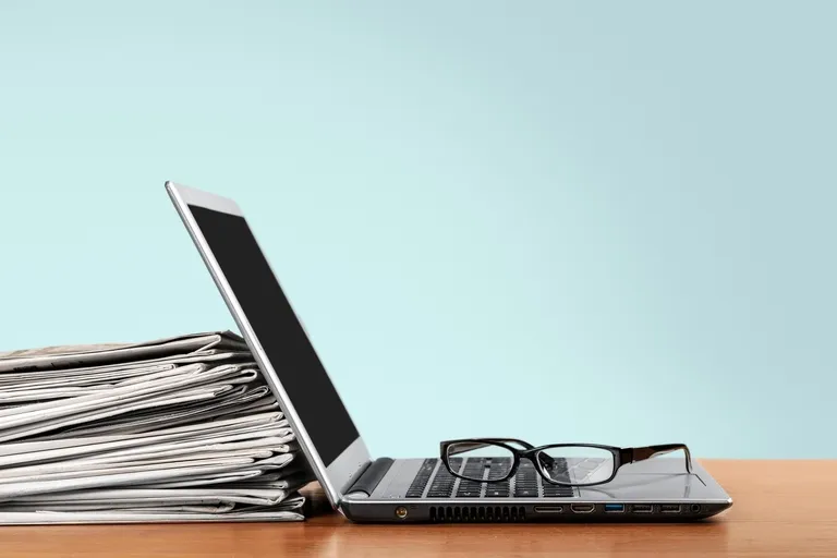 Stack of newspapers beside open silver laptop with black glasses on keyboard.