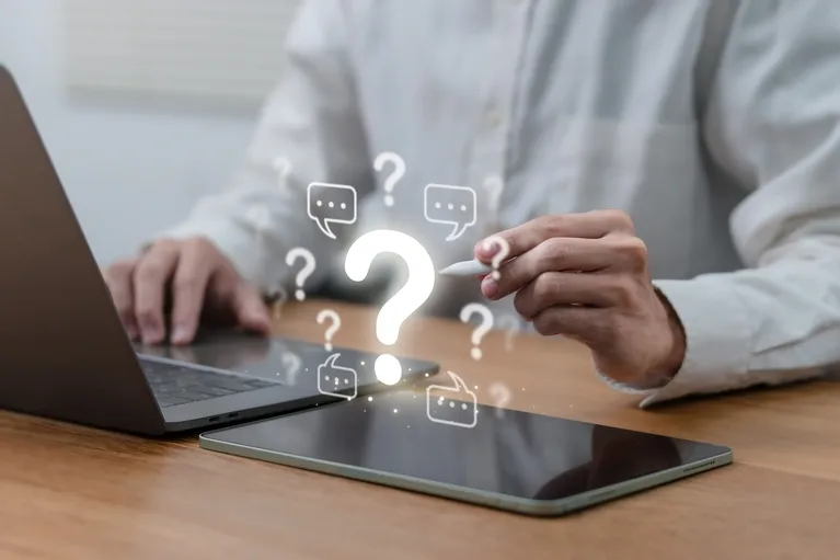 Person using stylus near tablet and laptop with floating question marks and chat icons.
