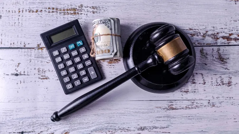 Black judge's gavel, calculator, and rolled US dollar bills on white wooden surface.