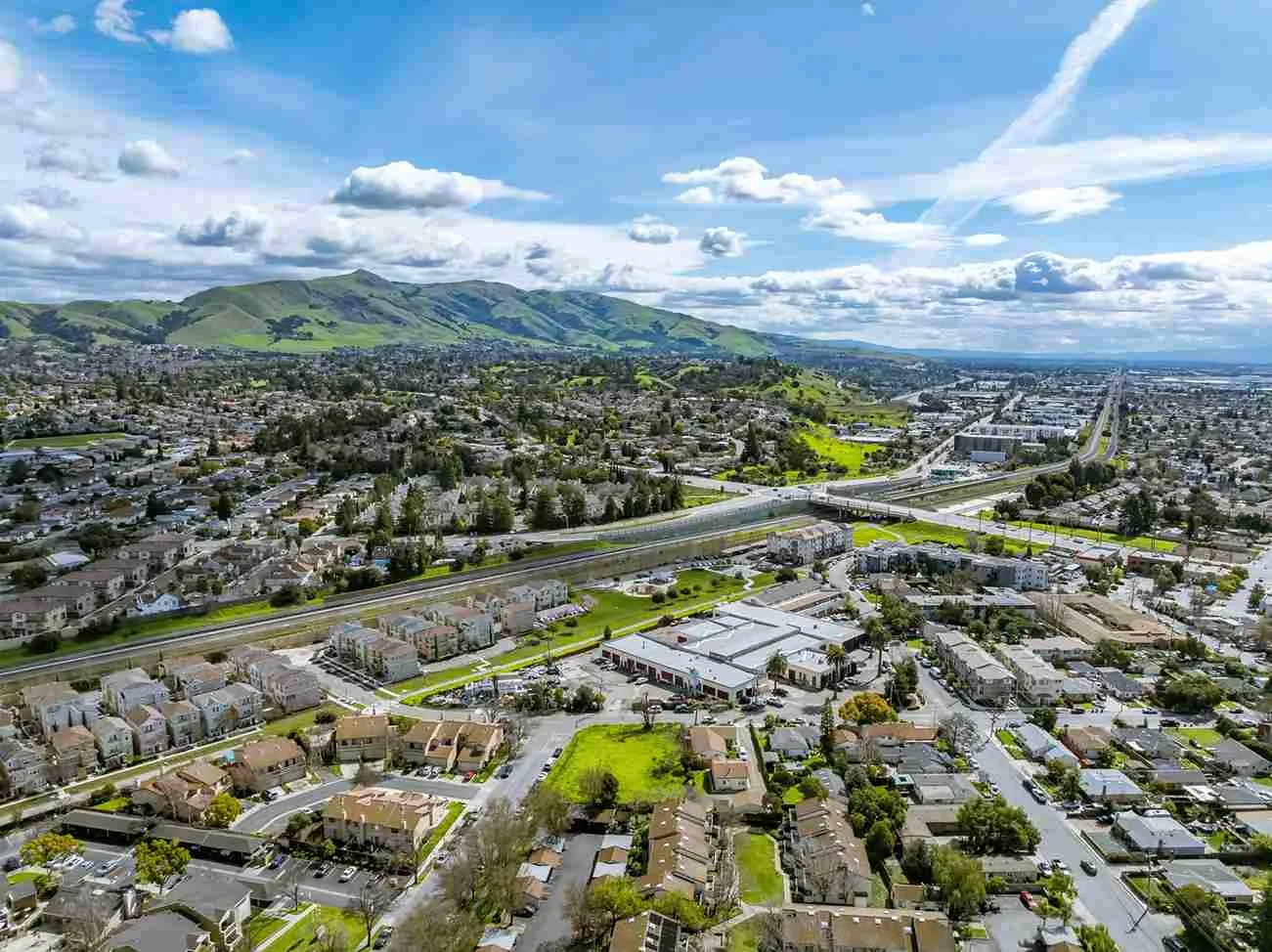 Aerial view of a suburban neighborhood with residential buildings, roads, highways, and green hills under a partly cloudy blue sky.