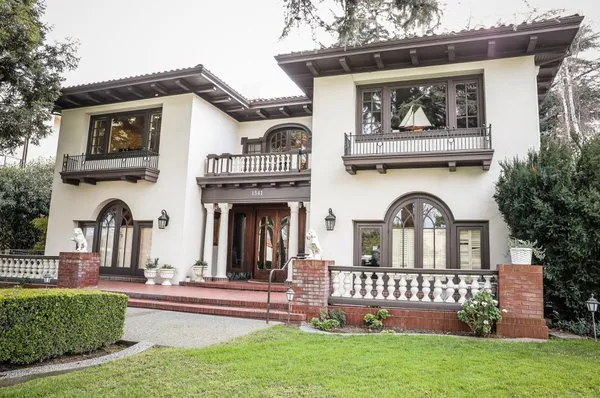 Two-story beige stucco house with dark brown trim, arched windows, balconies, and manicured lawn.