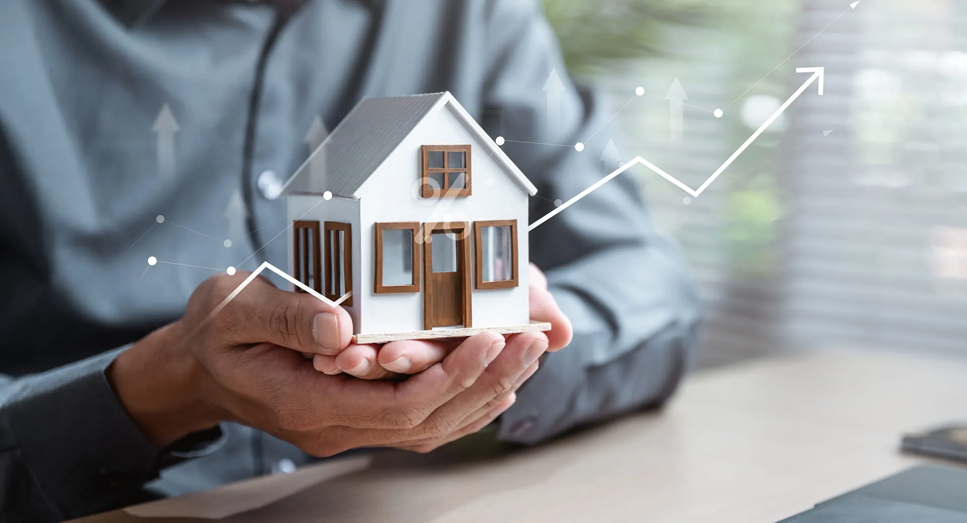 Person in gray shirt holding small white model house with digital upward graph overlay.