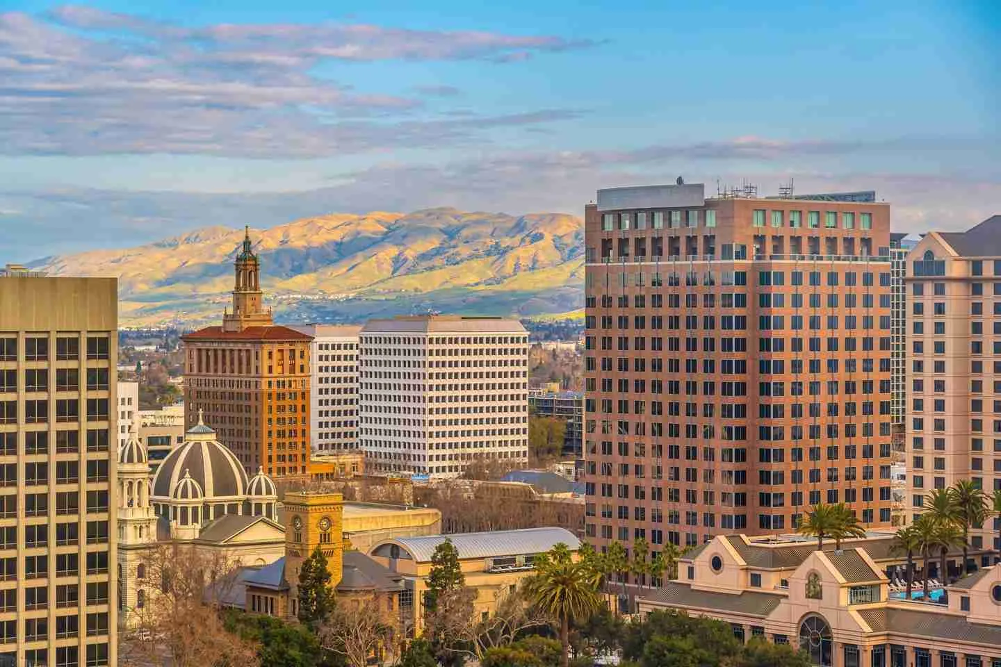 Cityscape featuring modern and historic buildings with mountains in the background under a partly cloudy sky.