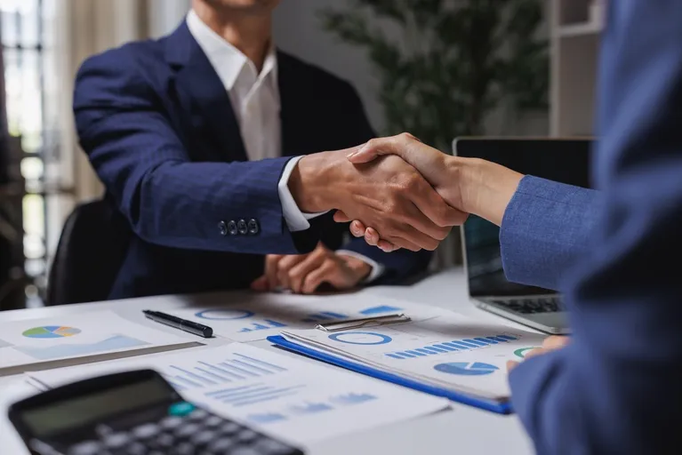 Two business professionals in blue suits shaking hands over a desk with financial charts and a calculator.
