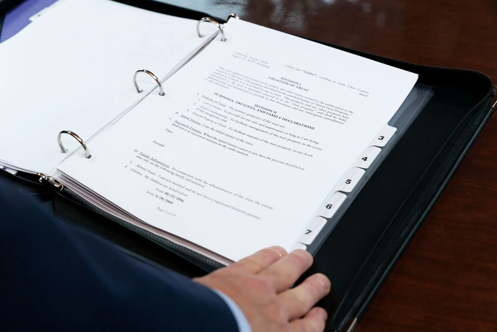 Open black binder with white tabbed documents, a hand turning a page on a wooden desk.