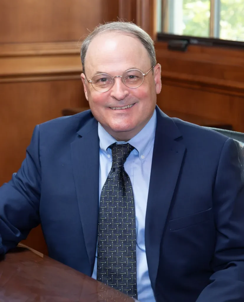 Middle-aged man in navy suit and patterned tie, seated in wood-paneled office.